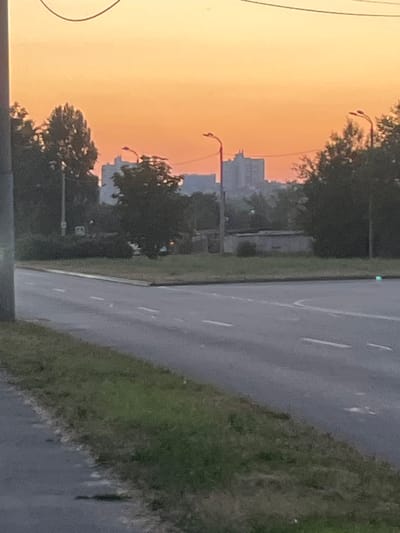 The image depicts a street scene at dusk in Dnipro, Ukraine. The sky is a hazy orange, suggesting either sunset or pollution. On the left side of the frame, silhouetted trees and a few tall buildings are visible, indicating an urban or suburban setting. Two streetlights with curved arms are present, one higher than the other. The road, on the right side of the image, appears wet, possibly from recent rain. Green grass lines the side of the road. There are no people visible in the image. The overall mood is quiet and slightly melancholic, with the fading light and muted colors. No text is visible.