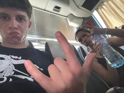 Two individuals are depicted in a selfie-style image. A female, holding a water bottle, is visible in the left portion of the frame making a hand gesture. To her right is a red toy car. A male is to the right, wearing a black t-shirt with a white design, also making a hand gesture. The background suggests an interior setting with window blinds visible. The location cannot be identified from the information given.