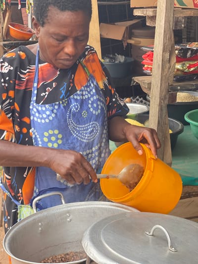 The image shows a woman engaged in food preparation within what appears to be a market stall or an informal kitchen setting. The image is captured from a slightly elevated perspective, looking down at the woman and her activity.
The central figure is a woman with dark skin and short, dark hair. She is wearing a patterned dress or apron with a blue upper section featuring white paisley and dot patterns, and a lower section with vibrant orange, black, and white abstract or floral designs. Her expression is focused, and her eyes appear to be looking down at her task.
She is holding a large, bright orange plastic bucket in her left hand, which contains a brown, stew-like food. A large metal spoon is immersed in the food within the bucket, held by her right hand. A faint wisp of steam is visible rising from the food in the bucket, suggesting it is warm or hot.
To the right side of the frame, a large silver aluminum pot is visible. The pot has a matching lid that is partially lifted or resting on its edge, revealing light-colored beans or peas inside the pot. The woman's hands and the bucket are positioned above and slightly to the left of this large pot, suggesting she might be about to transfer food into it, or serving from it.
The background reveals more of the setting. There are rough wooden planks or shelves behind the woman, supporting various items. These include packaged goods in shiny sachets or bags (some red, some black), and other containers like green plastic tubs. The overall environment suggests a bustling, functional space, possibly a roadside food vendor or an indoor market stall, characterized by practical, accessible materials and storage. The lighting appears to be natural and bright, indicating it is daytime.