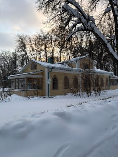 This image captures a snow-covered building in posёlok dachnogo khozyaystva «Arkhangel'skoe», Russia, under overcast winter conditions. The primary subject is a two-story, pale yellow structure with a dark, snow-laden roof from which multiple icicles hang. A green gutter system is visible along the roofline. The building features an enclosed veranda with multiple rectangular windows on its left side, displaying internal illumination. The main facade presents several arched, Gothic-style windows. Bare shrubs protrude from the deep, undisturbed snow covering the foreground and surrounding ground. In the background, numerous deciduous trees with bare, dark branches, some also laden with snow, are silhouetted against a cloudy sky that shows a warm, diffused glow near the horizon. No individuals or activities are depicted.