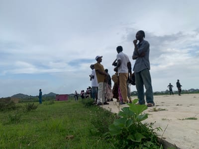 A group of people stand in a line on a paved surface, with a grassy field to their left and a sky with scattered clouds above. In the background, there are several structures, including a small pink building, and rolling hills. The people appear to be mostly young men, dressed in casual clothing. Some are looking out at the distance, while others are engaged in conversation. One man in the foreground, wearing a checkered shirt and jeans, has his arm crossed and is looking thoughtfully into the distance. The overall scene suggests a gathering or waiting period. The time of day appears to be daytime, possibly afternoon, given the lighting. The weather seems to be overcast but not raining. The location cues, such as the architecture of the buildings and the vegetation, point to a rural or semi-rural setting in Nigeria. No visible text is present in the image.