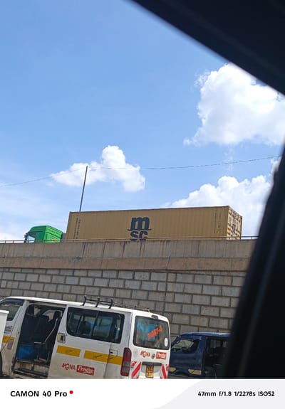 The image depicts a daytime scene in Nakuru, Kenya, observed from within a vehicle. Above, a bright blue sky with scattered white cumulus clouds is visible. A power line traverses the mid-ground horizontally. Directly below the sky, an elevated concrete retaining wall, constructed from large, light-colored blocks, supports a roadway. On this upper roadway, a yellow shipping container, marked with the black "MSC" logo, is being transported; only the green cab of the transporting truck is visible on the far left. Below the elevated structure, on a lower road, traffic is present. A white Toyota Hiace-style passenger van (matatu) occupies the lower-left foreground, its side sliding door open to reveal interior seating. The van features "MONA Prestige" branding, "BIGGY-G" on its rear window, a roof rack, and a partially visible Kenyan license plate "KBA 063Y" over red and white chevron hazard markings. To the right of the white van, a dark blue vehicle is partially visible. A dark, out-of-focus diagonal element frames the image from the upper right, suggesting the photograph was taken from inside a vehicle.