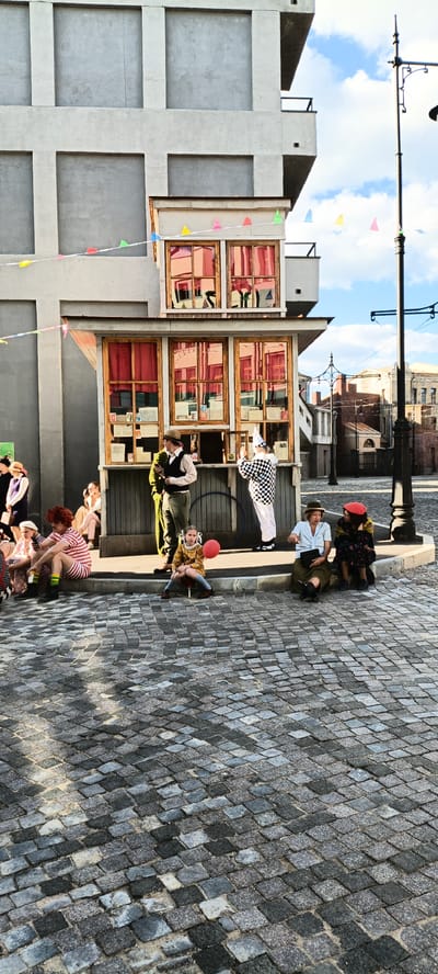 This media file captures a scene with several people dressed in vintage attire, gathered around a small, old-fashioned kiosk or shop. The setting appears to be a street with cobblestone pavement and a building with a modern, unfinished facade in the background, contrasting with the vintage elements. Colorful triangular flags are strung across the scene, adding a festive touch.

The subjects include a group of individuals in period costumes, suggesting a performance or reenactment. One person in a harlequin costume stands near the kiosk, possibly an employee or performer. Others are seated on the curb or the cobblestones, some appearing to be spectators. A young girl with a red balloon sits prominently in the foreground, her expression neutral. Another person with bright red, curly hair is dressed in a striped outfit and also sits on the curb.

The activity appears to be a gathering or a theatrical performance set in an earlier era. The kiosk itself has several display windows showcasing what look like old books or newspapers. The overall mood is relaxed and somewhat theatrical.

Notable details include the cobblestone street, the modern building construction in the background, and the vintage clothing of the people. The time of day seems to be late afternoon or early evening, indicated by the warm lighting and shadows. The weather appears to be clear. There is no visible text in the image. The location context is Moskva, Russia.