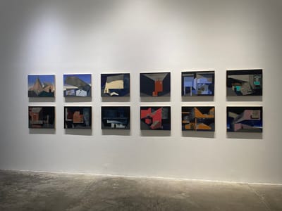 The image shows a white-walled room, likely a gallery, featuring a display of ten small, square, abstract paintings hung in two rows. The paintings have a dark, muted color palette with geometric shapes. The foreground displays a light-gray concrete floor. There are no people in the image. The location may be in Mexico City, Mexico.