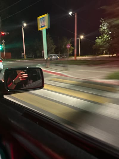 The image shows a night view from inside a car, looking into the side mirror and out the window. The car appears to be in motion, as evidenced by the motion blur in the image. In the side mirror, a person's hand, holding a smartphone, is visible, reflected from inside the car. The scene outside the window depicts a street at night, with a pedestrian crossing clearly visible in the foreground. Streetlights illuminate the area, casting light on the road and surrounding trees. Traffic lights are also visible, indicating an intersection. In the background, there are buildings and other cars. The location is identified as the city of Kostanay, Kazakhstan.