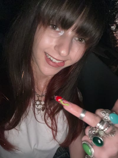 A woman poses closely, tilting her head to the left, displaying a smile. She has brown hair with bangs and light eyes. She wears a white shirt and multiple silver rings on her right hand, including those with turquoise, green, and light-colored stones. Her right index finger points downward, adorned with a red nail featuring a green worm design. A silver necklace with a Star of David charm hangs around her neck.