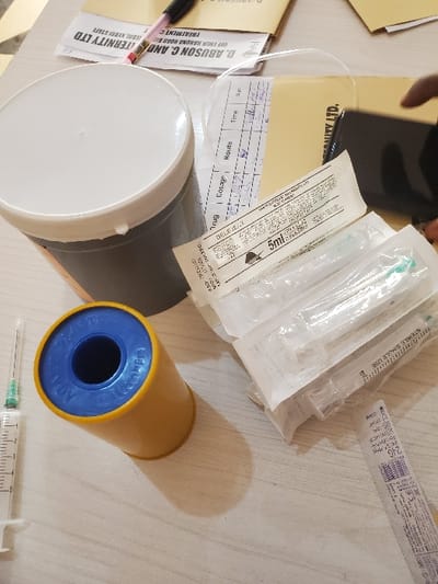 The scene depicts a medical preparation area on a light-colored surface, likely a table, within or associated with D. Abuson C. and Maternity Ltd., located in Birnin Kebbi, Kebbi State, Nigeria.
Forensically, the following objects are arranged:
A grey cylindrical container with a white lid is positioned in the upper left quadrant. Below it, a yellow cylindrical sharps container with a blue opening, labeled "ADVANCE MEDICAL," stands upright. To the immediate left, a single unpackaged 5ml syringe with a green needle cap is horizontally oriented.
Centrally, a stack of individually packaged sterile 5ml syringes, some marked "DELFLEX," are present; one package indicates a "24G" needle. Behind these syringes lies a white medical administration record form, containing columns for "Drug," "Dosage," "Route," "Time," and "Sign," with handwritten entries.
In the upper background, several manila envelopes or folders are visible, along with various loose documents, some of which bear the text "D. ABUSON C. AND MATERNITY LTD." A pink and black pen rests on one of these documents. On the far right, a partial view of a dark-skinned hand is seen near a dark-colored mobile phone and additional papers. A small white strip of paper displaying "24G" and barcode-like markings is located in the bottom right corner. The arrangement suggests an active setup for medical procedures involving injections or medication administration.