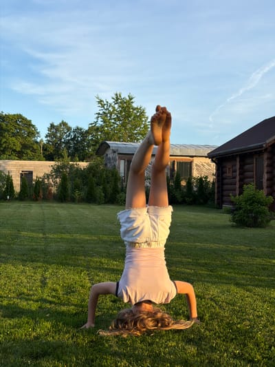 In the city of Dreiliņi, Latvia, a young person is performing a headstand on a lush green lawn. They are wearing a light pink t-shirt with a dark trim and white denim shorts. Their legs are extended upwards, and their bare feet are visible against a bright blue sky with wispy clouds. The background features a rustic wooden cabin with a dark brown roof, a small building with weathered siding, a low concrete wall, and several evergreen trees. The setting appears to be a backyard or rural property, with the warm glow of sunset casting long shadows across the grass.