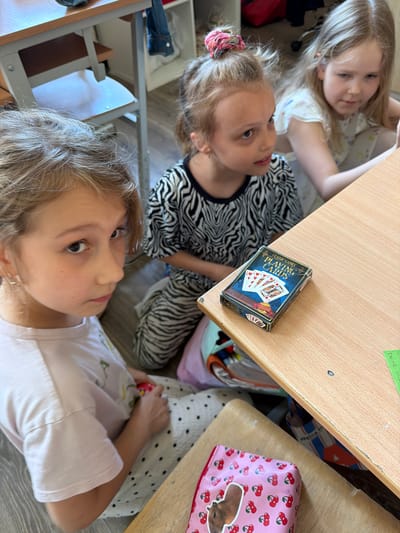 This image depicts three young girls in an indoor setting, likely a classroom or a similar educational environment, given the presence of desks and what appears to be a shelf unit in the background. One girl in the foreground, with blonde hair tied up with a pink scrunchie, is wearing a black and white zebra-print top and is seated on the floor. Her gaze is directed towards the viewer. To her right, another girl with long blonde hair, wearing a white top with a floral pattern, is also seated and looking towards something to her right, possibly off-camera. The third girl, whose face is only partially visible in the foreground, has blonde hair and is wearing a light pink t-shirt. She is looking towards the viewer and her left.

On a light-colored wooden desk in the middle ground, a deck of "Classic Games Playing Cards" is placed. The card box is dark blue with an illustration of playing cards. There are also some papers and bags visible on and around the desk. The overall atmosphere suggests a casual gathering or activity among the children. The image does not provide any visual cues that would allow for the determination of the specific location within the city of Riga, Latvia.