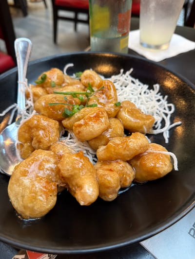 This image captures a close-up, top-down view of an appetizing dish served in a glossy black ceramic bowl, likely in a restaurant setting in Puebla, Mexico. The main subject is a generous portion of golden-brown, battered pieces of food, possibly shrimp or chicken, coated in a glistening, translucent light brown sauce that suggests a sweet and savory profile.
Beneath the main protein, a bed of finely shredded, crispy white strands, resembling fried rice vermicelli or thin noodles, provides a textural contrast. The dish is garnished with finely chopped green herbs, likely chives or green onions, adding a touch of freshness. A silver spoon is visible inside the bowl, resting among the food.
In the blurred background to the left, a tall, frosted glass containing a pale yellow liquid, possibly lemonade or a cocktail, stands beside a colorful bottle with red, green, and yellow bands. The dark table surface and indistinct shapes suggest an indoor dining environment. A small, partially visible piece of paper or card to the right of the bowl shows the upper part of the letter "P." No people are visible in the frame. The lighting is bright and direct, highlighting the inviting appearance of the meal.