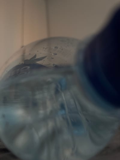 The media file is a close-up, low-angle shot of the neck and upper portion of a clear plastic bottle, likely containing a light-colored liquid, possibly water. The bottle is filled with the liquid, and small bubbles are visible within it, particularly towards the right side of the frame. A dark blue cap or lid is visible at the very bottom of the image, obscuring part of the bottle's opening. Inside the bottle, towards the upper right quadrant, a dark, abstract shape resembling a stylized leaf or feather is discernible, possibly part of a label or design. The background is out of focus and appears to be a light-colored, neutral surface, possibly a wall or a piece of furniture, with subtle horizontal lines suggesting paneling or blinds. The lighting is soft and diffused, creating a slightly hazy or ethereal effect, and there are no discernible human figures or animals. The overall impression is one of stillness and simplicity, with the focus being entirely on the bottle and its contents. No specific time of day or weather conditions can be inferred. The location is unknown.