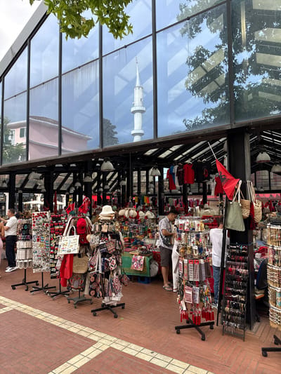 This image depicts a bustling outdoor market scene, likely situated within a covered walkway or under a large glass roof structure. The ground is paved with reddish-brown bricks, featuring decorative lighter-colored stripes.
Numerous market stalls, laden with a wide variety of merchandise, line the right side of the frame. These stalls display an assortment of goods including hats (straw, red, and white styles), various bags (woven, patterned fabric, and handbags), and clothing items such as t-shirts. Some t-shirts are visibly printed with the word "ALBANIA" and a double-headed eagle motif. Also prominent are numerous tourist souvenirs like keychains, magnets, and a diverse collection of jewelry, including bracelets, necklaces, and pendants, with many featuring evil eye charms. A distinctive red Albanian flag, adorned with its black double-headed eagle, is proudly displayed on a pole above one of the stalls, serving as a clear national identifier.
Several individuals are present, engaged in market activities. A vendor, wearing glasses and a patterned shirt, sits behind a stall, observing the scene. Other people, partially visible, appear to be browsing the merchandise. One person is seen from the waist down, wearing a white t-shirt, dark shorts, and flip-flops, standing at a stall.
A striking feature of the scene is the large glass ceiling above, which reflects the surroundings. These reflections show a bright, clear sky, green foliage from nearby trees, and significant architectural elements. Most notably, a tall, white minaret-like structure, characteristic of a mosque or a clock tower in Tirana, is clearly reflected, providing a strong geographical cue. Other buildings are also faintly visible in the glass.
The overall impression is of a vibrant market operating on a bright, clear day, suggesting good weather conditions suitable for outdoor shopping. The atmosphere appears casual and lively, typical of a busy local or tourist attraction.