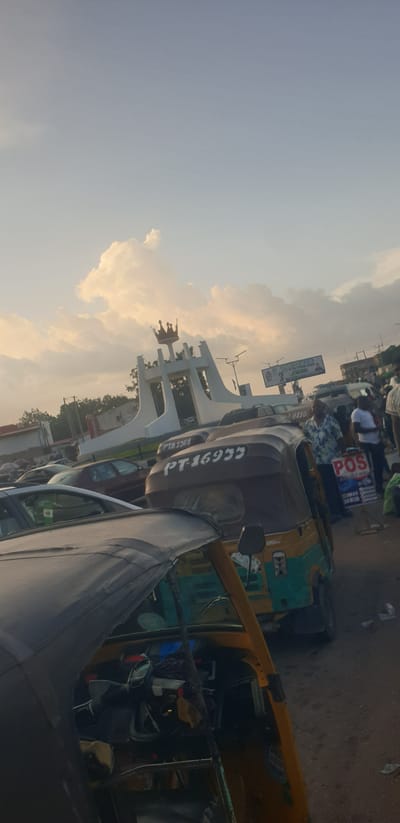 This media file shows a street scene in Barakin Lerawi, Nigeria, at what appears to be late afternoon or early evening. The sky is a pale blue with some fluffy, golden-tinged clouds, suggesting a warm and pleasant atmosphere. The scene is dominated by several auto-rickshaws, also known as Keke Napeps, which are common modes of transportation in Nigeria.
One auto-rickshaw in the foreground is primarily gray, with a black tarp covering part of it. Another in the mid-ground is a mix of dark brown and green, with yellow accents. A third auto-rickshaw, visible further back, is predominantly yellow. People are visible in and around the auto-rickshaws, suggesting they are either passengers or drivers. Some individuals are wearing casual clothing.
In the background, there is a large white monument with intricate designs, possibly a landmark or an architectural feature. A billboard or sign with text is also visible above the monument, and a smaller sign with "POS" is prominent on a blue background, indicating point-of-sale services. The road surface appears to be dirt or gravel. The overall impression is of a busy, active street with the hustle and bustle of daily life. The sunlight casts a soft glow on the scene, creating a warm ambiance. The orientation of the image is rotated, with the sky appearing on the left side of the frame.