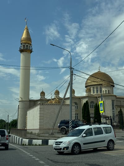 The image depicts a street scene in Ust-Dzheguta, Russia, prominently featuring a mosque with a tall, slender minaret topped with a golden, conical roof adorned with a crescent moon. The main mosque building has a large, golden dome also decorated with a crescent moon, and several smaller golden domes. The architecture is light-colored stone. In the foreground, a white station wagon is driving on a road with a pedestrian crossing. Other vehicles, including a dark SUV and another silver SUV, are visible. The environment includes trees, streetlights, power lines, and a clear blue sky with scattered clouds. A yellow pedestrian crossing sign is visible near the mosque entrance. The overall scene suggests a sunny day in a city.