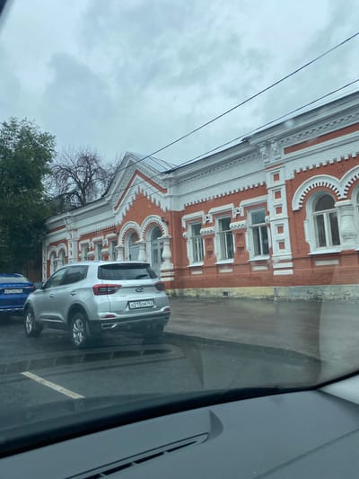 The image shows a silver Chery Tiggo 4 Pro car parked on a wet asphalt street in front of a red brick building with white ornate trim and arched windows. The building appears to be a historical or architecturally significant structure. A blue car with a license plate is partially visible to the left of the silver car. The sky is overcast and grey. The photo is taken from inside another vehicle, looking out through the windshield. The license plate of the silver car reads H215OM 763. The license plate of the blue car reads E572BH 763. The scene is likely in Novokuybyshevsk, Russia, given the style of the license plates and the architecture.