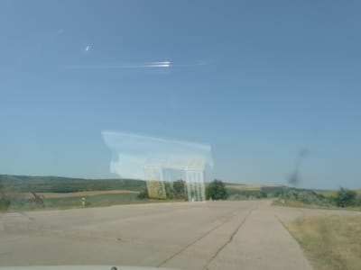 The image shows a roadside scene in Glinjeni, Moldova. A pack of cigarettes, with the brand name "CYMBER" visible, is positioned at an angle on the side of a paved road. The road appears to be a rural highway with fields and trees in the background. The sky is clear and blue. The image is taken from inside a vehicle, as evidenced by reflections on the surface. There are no people or animals visible in the frame. The overall environment suggests a sunny day in a rural area.
