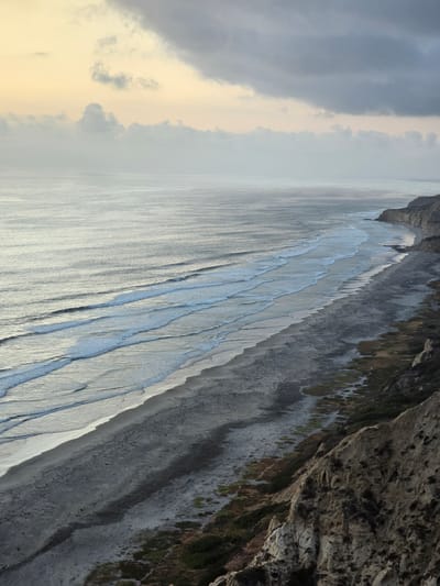 This is an aerial view of Torrey Pines State Beach in San Diego, California, on a cloudy day. The ocean stretches to the horizon, with gentle waves rolling onto the sandy shore. The beach is wide and appears mostly deserted, with a few scattered individuals walking along the waterline. To the right, the dramatic cliffs of Torrey Pines rise, covered in sparse vegetation. The sky above is overcast, with patches of pale yellow and gray clouds, suggesting either sunrise or sunset. The overall mood is serene and natural.