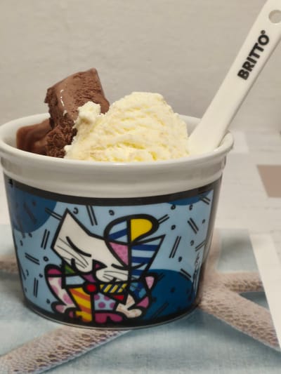 The image shows a close-up of a cup filled with ice cream and a Britto branded spoon. The cup is decorated with a colorful, abstract cat design. The ice cream appears to be chocolate and vanilla flavors. The background is a light-colored textured surface, possibly a table. The image is taken indoors and there is no visible indication of the location being Passignano sul Trasimeno, Italy.
