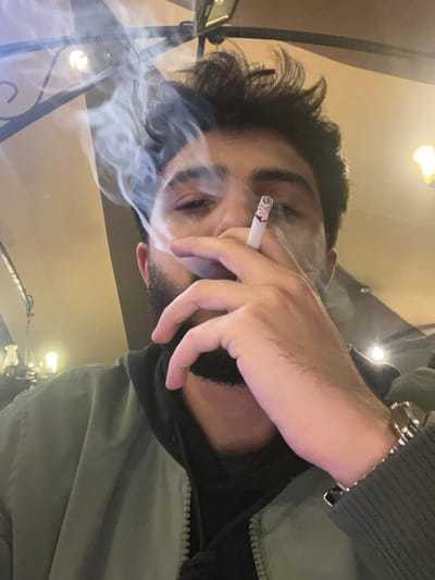 A man smokes a cigarette indoors. White smoke fills the frame around his face and obscures the background, which includes visible light fixtures. He wears a green jacket over a black hoodie. A silver wristwatch is visible on his left wrist. His dark hair is messy. He looks down slightly, with a pensive expression.