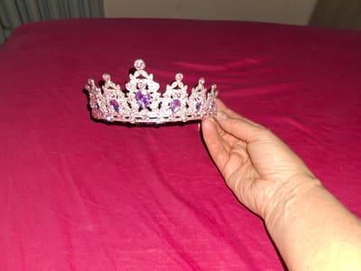 A hand, likely belonging to a person, is holding a delicate, ornate tiara adorned with pink and purple gemstones against a vibrant magenta background. The tiara features intricate silver-toned metalwork with dangling teardrop-shaped pink stones and larger, faceted purple stones at its center. The hand's skin is light-toned with visible veins and creases, suggesting it belongs to an adult. The magenta fabric behind the tiara appears to be a textured material, possibly clothing or upholstery, and is slightly wrinkled, indicating it is draped or folded. The lighting in the image is somewhat dim, casting subtle shadows around the tiara and the hand. The focus is sharp on the tiara and the hand, while the background is softly blurred. There is no visible text in the image, and no specific cues to suggest the time of day, weather, or a particular location beyond the provided context of Ogre, Latvia. The overall impression is of a close-up shot of someone presenting or showcasing the tiara.