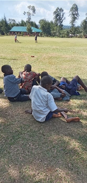 The image shows five young boys sitting on a grassy field. The boys are all wearing casual clothing. The boy on the right is wearing a white long-sleeved shirt. The boy in the center is wearing a red and white patterned shirt. The boy on the left is wearing a blue shirt and pants. The boys are sitting in a circle and seem to be talking to each other. The scene is set in Bungoma, Kenya, with the coordinates (latitude: 0.553, longitude: 34.773). There is a small, light-blue roofed building in the background. The field is surrounded by trees and appears to be in a rural setting.