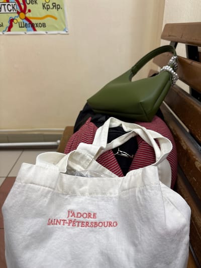 A green purse with a pearl handle, a red and white striped pillow, a black garment, and a white tote bag with the text "J'ADORE SAINT-PÉTERSBOURG" in red embroidery hang from a wooden coat rack. A poster with Cyrillic text and what appears to be a map is partially visible on the left. The location is Ust-Ilimsk, Russia, as indicated by the Cyrillic text. The objects are likely personal belongings placed on the coat rack. The environment is indoors with tiled flooring.