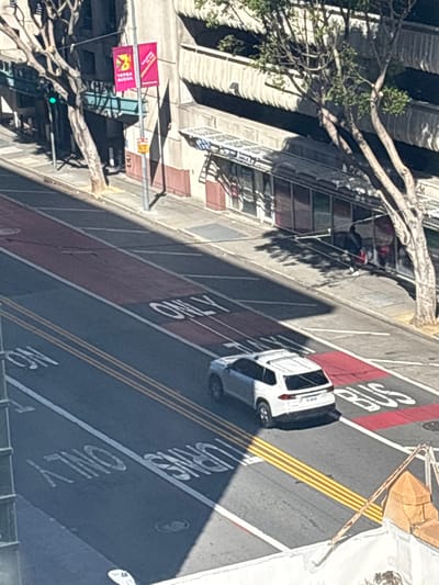 This image, taken from a high vantage point in San Francisco, California, depicts an urban street scene. A white SUV is captured in motion on the right side of the frame, navigating a lane marked with yellow and white stripes. Adjacent to this lane, a red-painted section of the road bears the white stencil "BUS ONLY." Further to the left, another lane is marked with the words "ONLY TURNS" and "ONLY," indicating specific traffic regulations.
In the background, the architecture of the city is visible. Buildings with varying facades line the street, some featuring awnings and windows. A streetlamp with a green traffic signal is present on the left side of the image, alongside a tree with bare branches. Two pink banners hang from a pole, one displaying "VERNA BUENA" and the other an illegible message. The presence of pedestrians is noted in the background, near the bus stop area. Shadows cast by trees and structures suggest it is daytime, with the sun positioned to the left. The overall impression is a typical street view in a bustling city environment, highlighting traffic management elements and urban infrastructure.