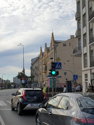 This is a photograph taken from a street-level perspective, with the image rotated 90 degrees counter-clockwise. The scene depicts a city street with buildings on either side and cars on the road.

In the foreground, the back of a dark-colored car, identified as a Toyota Aygo, is prominently visible. Its license plate reads "WWL 569PR". To the right of this car, a portion of a larger, dark grey car is visible.

On the street, a traffic light is illuminated with a green signal. Adjacent to the traffic light are several road signs, including a yellow diamond-shaped sign and a blue sign with a pedestrian symbol. Further down the street, more traffic lights and signs can be seen.

The buildings lining the street appear to be older, with ornate architectural details, especially on the yellow building to the left. Some buildings have balconies and intricate window frames. Graffiti is visible on some of the building walls, suggesting an urban environment.

Several people are visible in the background, mostly standing on sidewalks or near the buildings. They appear to be dressed casually. One person is wearing a bright yellow vest.

The sky is filled with white, wispy clouds against a pale blue background, indicating daytime. The overall impression is a bustling city street in Poland, possibly Warsaw, during fair weather. The time of day appears to be either late morning or afternoon.