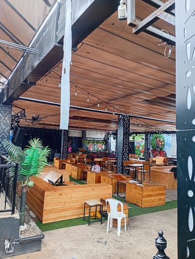 This media file captures an outdoor seating area of a restaurant or cafe. The scene is filled with rows of wooden tables and benches, some of which appear to be communal seating. A white plastic chair is visible in the foreground. The area is covered by a wooden roof structure, and string lights are hung overhead. Green artificial turf is laid out between some of the seating arrangements. In the background, there are posters or artwork displayed on the walls, including one with a wreath and a person wearing red, and another with a tropical theme. There are also glimpses of what appears to be sound equipment, including speakers and a fan.
The image is taken from a high angle, looking down onto the seating area. The lighting suggests it could be daytime, though the covered nature of the seating area makes it difficult to ascertain the exact time of day or the weather conditions. The presence of multiple tables and chairs suggests a place open for business, likely a cafe or restaurant. The overall impression is a casual, outdoor dining or gathering space. The provided location context of "Howayi, Nigeria" suggests this is a business operating within that region. No specific activity or event is clearly depicted, but the setup implies it's a place for patrons to relax and dine.
