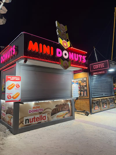 The image depicts a nighttime scene at an amusement park or fairground, possibly in Nesebar, Bulgaria, as suggested by the context. Several food stalls are visible, illuminated by neon and digital signage. A prominent stall features a large neon sign reading "MINI DONUTS" in pink and red letters, with an illuminated donut logo above. To its right, another stall advertises "COFFEE" and "CREPES & WAFFLES" with similar signage.

In the foreground, a smaller stall displays images of hot dogs with prices on a sign that reads "THE AMERICAN HOT DOG". The stall itself has a window displaying promotional imagery for "MINI DONUTS" with the "Nutella" brand. The ground is paved with cobblestones and sand, suggesting an outdoor environment. A partially visible Ferris wheel can be seen in the upper left corner, further indicating an amusement park setting. A black electric scooter is parked near the waffle stall. The overall atmosphere is lively and commercial, typical of a nighttime entertainment venue.