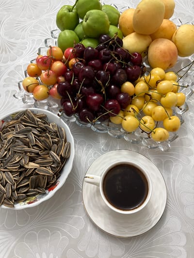 A close-up, top-down shot features a white cup of dark coffee on a saucer, positioned to the right of a white ceramic bowl filled with sunflower seeds. To the left of the bowl is a tiered glass fruit platter overflowing with various fruits, including green plums, apricots, dark red cherries, and yellow cherries. The arrangement is set on a white tablecloth with a subtle embossed floral pattern. The image appears to be taken indoors, as suggested by the lighting and the indoor setting. There are no people or animals visible in the scene. The overall impression is that of a simple, healthy snack or breakfast spread.