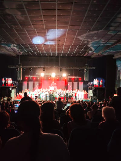 The image depicts a large indoor gathering, likely a performance or event, with an audience seated in a darkened venue facing a brightly lit stage. The stage features numerous performers, many in colorful costumes, arranged in a semi-circle. The ceiling of the venue is visible, featuring a grid-like structure with projected images of clouds and stars, suggesting a decorative or thematic element. The audience members in the foreground are largely silhouetted, with some individuals holding up phones, indicating they may be recording or photographing the event. There is a noticeable absence of specific landmarks or identifiable features that would allow for precise location identification within the city of Mykolaiv, Ukraine. The overall scene conveys a sense of communal engagement with a live performance in a theater or similar venue.