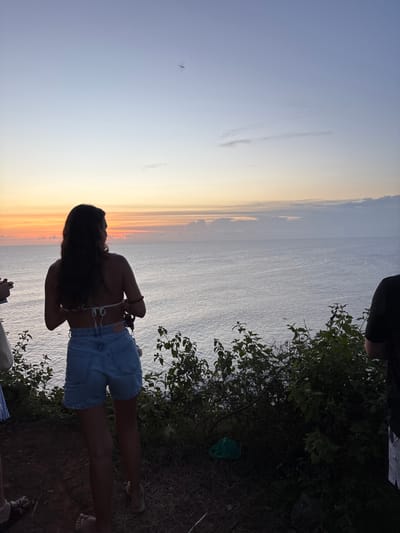 Captured in Pecatu, Indonesia, the image presents a twilight scene where a woman stands silhouetted against a horizon transitioning from a vivid orange sunset to a muted blue sky above a body of water, presumably the ocean. The woman, clad in denim shorts and a light-colored top, is positioned near the edge of vegetation, with glimpses of plant life on her right. In the distance, a small aircraft is visible in the sky. The overall composition suggests a viewpoint from a cliff or elevated location.