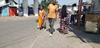 On a sunny day, three individuals walk on a paved road near buildings. A woman in an orange patterned dress walks alongside a man in a brown t-shirt and dark pants. A woman with long black hair in a patterned blue dress walks to the right. Behind them, a building with gas pumps is visible. Further to the right, men stand near a wooden structure.