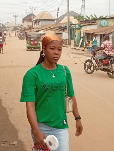 An outdoor scene in Umuode, Nigeria, depicts a woman in the foreground walking on an unpaved, reddish-brown dirt road. She wears a golden-brown headscarf, a green short-sleeved t-shirt with dark lettering, light blue jeans, silver-colored dangle earrings, and a necklace with a pendant. A light-colored shoulder bag with a chain strap is on her left shoulder, and a dark wristwatch is on her left wrist. She holds a white portable fan and a striped object in her right hand.
In the midground, a green and red motorized tricycle (Keke Napep) is visible moving away on the left side of the road, behind a walking man in a grey t-shirt and red shorts. On the right, a man on a red motorcycle with chrome accents is positioned towards the foreground. Buildings with various roof types, including corrugated metal and flat, line both sides of the road. Multiple electrical poles with overhead wires crisscross the scene. Satellite dishes are mounted on some buildings. A tall metal lattice structure is visible in the far background on the right. The sky is overcast.