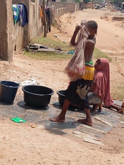 The image depicts an outdoor scene in Lafia, Nigeria, characterized by an unpaved, sandy environment under bright sunlight. In the foreground, a barefoot individual with dark skin, wearing yellow shorts and dark trousers, stands with their upper torso exposed, holding a wet, light-colored patterned garment aloft, appearing to wring or shake water from it. Adjacent to this individual, a second barefoot person with dark skin is seated on the sandy ground, wearing a maroon head covering (hijab) and a matching long garment.
Between and in front of the two individuals, three black plastic tubs are positioned on a section of wet, light-colored paving stones where water has pooled. A small green plastic item and a white bar of soap are visible on these wet stones near the tubs. The immediate ground is a mix of wet paving and dry, reddish-brown sand.
In the mid-ground on the left, a concrete wall has several colorful garments hanging to dry. The background reveals an unpaved dirt road extending into the distance, bordered on the right by a concrete wall with regularly spaced vertical pillars. Further in the background, residential structures are partially visible, and an area of debris or refuse is discernible on the right side of the road.