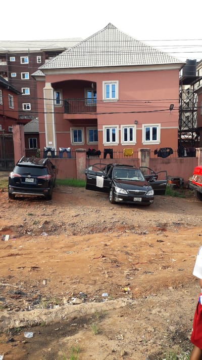 The image depicts a scene in Nkpor, Nigeria. A three-story salmon-colored building with white-framed windows is visible. Clothes are hanging on the balconies. Two black cars are parked in front of the building, one with an open door. A red vehicle is partially visible near the building. The ground is a mix of reddish-brown dirt, gravel, and trash. Power lines run in front of the building. Part of a person wearing a red shirt and a white shirt is visible in the top right corner.