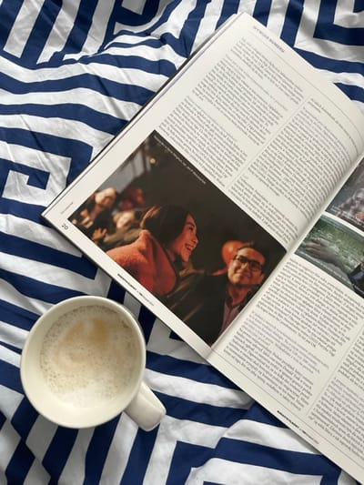An overhead view captures a domestic scene set in the city of Iława, Poland. A bed is covered with blue and white striped bedding, featuring a repeating geometric pattern. Resting on the bedding is an open print magazine, specifically "MAGAZYN FILMOWY" issue 188, dated November 2024, displaying page 20. The left page of the magazine features a prominent photograph of two individuals: a female with dark hair, wearing a reddish-brown garment, smiling broadly towards a male wearing glasses and a dark suit, who is also smiling. Additional blurry figures are visible in the background of this photograph. Accompanying text below the image identifies the subjects as "Natalia Buc w filmie Mignona, reż. Jan P. Matuszyński." The right page contains further Polish text. To the lower left of the magazine, a white ceramic mug, containing a light brown beverage topped with foam, is present. The overall arrangement suggests a paused leisure activity.