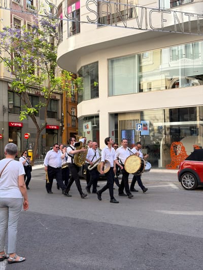 A street band is marching in a city street. The band members are wearing white shirts and dark pants. They are playing various instruments, including a tuba, a saxophone, a trumpet, and drums. The band is walking on the right side of the street, towards the viewer. In the background, there are buildings with shops on the ground floor. One of the shops has a sign that says "VIPS". There is also a red car parked on the side of the street. The street is paved with asphalt and there are some trees and streetlights. The sky is blue and there are some clouds. The image was taken in València, Spain, as indicated by the neon sign in the background that reads "SAN VICENTE". The overall scene suggests a festive event or parade.
