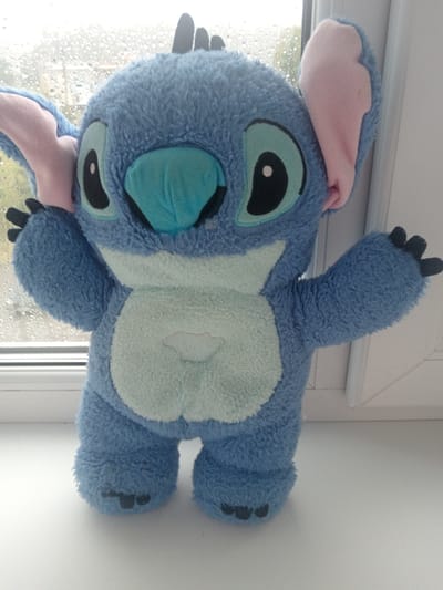 A plush toy resembling the character Stitch from Disney's Lilo & Stitch franchise stands upright on a white, flat surface. The toy is predominantly blue with lighter blue on its belly and a turquoise nose. It has large black eyes with light blue accents and pink inner ears. The toy's arms are outstretched to its sides. The background shows a window with raindrops on it, with a blurred outdoor scene visible beyond. The overall setting appears to be indoors, possibly on a windowsill. The location is identified as Кадзінскі сельскі Савет, Belarus, though this information is not visually verifiable from the image itself.