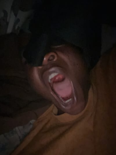 The image is a close-up, low-angle shot of a person's face with their mouth wide open, as if yawning or screaming. The lighting is dim, with the face illuminated from the front, creating shadows that obscure details. The person's eyes are visible, and their tongue can be seen inside their mouth. They appear to be wearing a light-colored t-shirt. The background is dark and out of focus, making it difficult to discern any specific environmental details. No specific location in Ikorodu, Nigeria can be identified from this image.