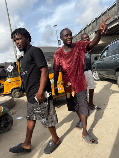Three Black men stand posing in front of an auto rickshaw in Lekki, Nigeria. One man in the foreground wears a black T-shirt and patterned shorts. He holds a white charger. The second man wears a maroon shirt and black shorts and is smiling. The third man is behind the second and is pointing upwards. The auto rickshaw is yellow and has writing on it. A car is parked behind them. Above them is an elevated road. The sky is cloudy.