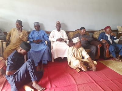 In the city of Barden Yana, Nigeria, a group of seven men are seated in what appears to be a reception or gathering. The men are dressed in traditional Islamic attire, consisting of flowing robes and caps. The seating arrangement suggests a formal occasion, with some men seated on chairs and others on the floor. The environment appears to be indoors, with a light-colored wall in the background and a red carpet covering the floor. One man in the foreground is seated on the floor, dressed in a dark blue outfit and black cap, with his legs crossed. He is looking down, and his hands are resting on his lap. To his right, another man is seated on a chair, wearing a light-colored robe and a cap, and appears to be speaking on a mobile phone. Next to him, a man in a blue robe and cap is seated, with his hands folded in his lap. Another man in a white robe and cap is seated in the center, with his hands clasped in front of him. Further to the right, two men are seated on a patterned sofa. One is wearing a dark gray robe and black cap, while the other is wearing a blue robe and a red cap. The overall impression is one of a calm and composed gathering.