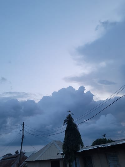 The image depicts a dramatic sky with heavy, dark clouds casting shadows. The scene is oriented vertically, with the sky dominating the left side and a partially visible structure on the right. This structure appears to be a building with a corrugated metal roof, possibly a house or a shop. Electrical wires are strung across the sky, some taut and others sagging, supported by a wooden pole visible in the lower right section. A tree with dense foliage is also visible, clinging to the side of the building, its branches reaching towards the sky.
The weather appears stormy or overcast, with the dense clouds suggesting impending rain or a recent downpour. The lighting is dim, indicative of a late afternoon or early evening, or perhaps the atmospheric conditions of a storm. The colors are muted, with shades of blue, gray, and white in the sky and the earthy tones of the building. The image evokes a sense of anticipation or the calm before a storm. There is no visible text in the image. The location context of Barakin Akawo, Nigeria, suggests an urban or semi-urban setting within that region.