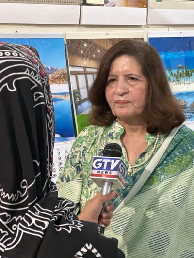 The image shows an older woman with brown hair being interviewed. She is wearing a green floral patterned top and a pearl necklace. A microphone with the "GTV News HD" logo is positioned in front of her. The interviewer is partially visible, wearing a black and white patterned garment. The backdrop includes several images: one of a tropical beach, another of an interior architectural space, and a calendar. The OCR indicates that the calendar is in Japanese, showing the numbers 2, 3, 9, 14, 15, 16, 22, 23, and 29. The location is Quetta City Tehsil, Pakistan.