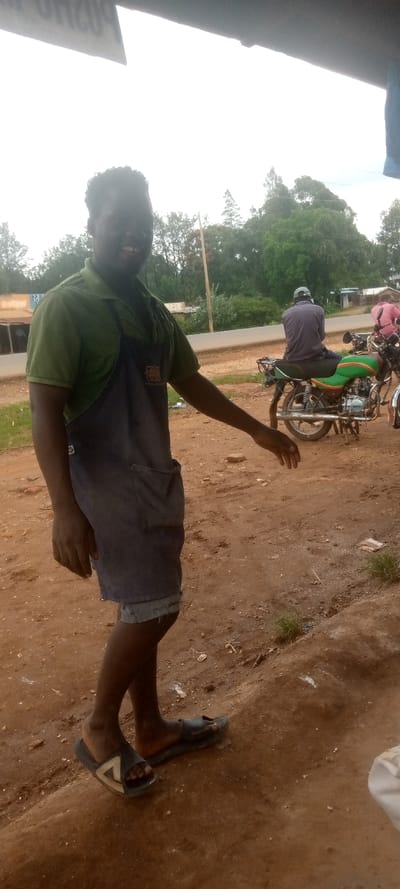 A young Black man wearing a green polo shirt, a blue apron, and black sandals is smiling and standing on a dirt path in front of a small store in Kivaywa, Kenya, near a motorcycle parked along the roadside.  He is facing the camera. Behind him are trees.  To the man's right, a Black man wearing a grey shirt and a black cap is sitting on a motorcycle, facing the opposite direction. The motorcycle is parked along the edge of the road.  There is also a third Black man standing on the opposite side of the road wearing a pink shirt.