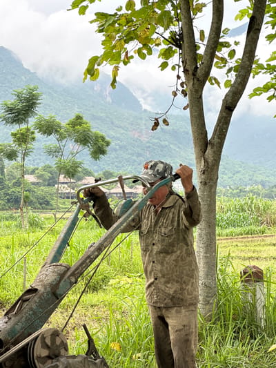 An older man in a camouflage cap with a red star and a camouflage patterned shirt stands in a grassy field, holding onto the handles of a green, weathered tiller. His pants are also stained with dirt. The man appears to be adjusting or operating the tiller, which is positioned in front of him. The background reveals a lush, green landscape with rice paddies, tall grasses, and trees. In the distance, mist-covered mountains rise, characteristic of the scenery in Mai Chau, Vietnam. The sky is overcast with clouds.