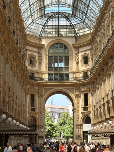 The image shows a view inside the Galleria Vittorio Emanuele II in Milan, Italy, looking towards the exterior through an archway. The architecture is ornate with classical columns and statues lining the walls. A glass and metal roof arches overhead, allowing natural light to filter into the enclosed space.
In the foreground, a crowd of people are visible, some walking and some gathered. The lower portion of the image shows the awnings of shops and restaurants. Brands like "Galleria" and "MONCLER" are partially visible on the awnings.
Through the central archway, a statue is visible in the distance, flanked by trees. Beyond the trees, buildings with classical architecture and a modern tower are present. The sky is clear and blue. The overall impression is of a busy, elegant urban space.