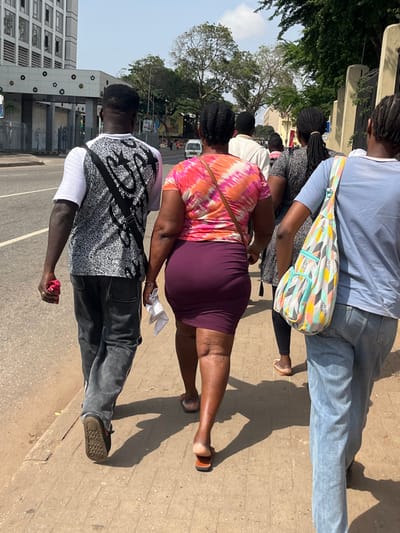The image depicts an outdoor urban scene in Accra, Ghana, specifically near the Accra International Conference Centre, identifiable by the distinctive building with circular and star-shaped facade patterns on the left.

Five individuals are observed walking away from the camera on a concrete sidewalk, adjacent to a paved road, under a clear, sunny sky.

From left to right in the foreground:
1.  A male individual wears a white t-shirt layered with a black and white patterned sleeveless top, dark jeans with white side stripes, and black closed-toe shoes. He carries a crumpled red item in his right hand and has a black bag strap diagonally across his back.
2.  A female individual wears a pink, orange, and white tie-dye short-sleeved t-shirt, a fitted plum-colored knee-length skirt, and open-toed brown sandals. A thin brown strap crosses her right shoulder, and she holds a folded white cloth in her left hand.
3.  A female individual wears a light blue t-shirt and light blue jeans. A multi-colored fabric bag with a geometric pattern hangs over her left shoulder.

Behind these individuals, two more individuals are partially visible:
4.  A female, mostly obscured, appears to wear a dark patterned dress or top and light-colored open-toed sandals.
5.  A male, mostly obscured, wears a light-colored long-sleeved shirt.

The sidewalk is concrete, and shadows of the individuals are cast upon it. A multi-lane asphalt road runs parallel to the sidewalk, with a white vehicle visible in the distance. Lush green trees and foliage line the right side of the road, contrasting with one tree appearing partially bare. The sky is bright and clear.