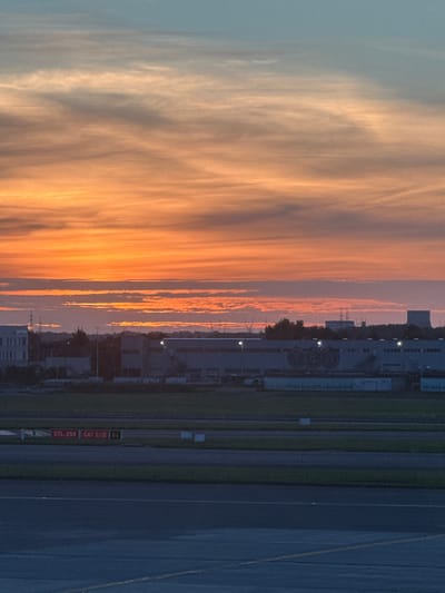 The image captures a sunset over Zaventem, Belgium, likely near Brussels Airport. The sky is ablaze with hues of orange and pink, transitioning into a soft blue. Below the sky, industrial buildings and infrastructure are visible. One building has a "SAFRAN" logo. Another building features a prominent advertisement with the text "we purify the airport's wastewater". The foreground shows a dark tarmac, possibly a runway or taxiway, with markings and caution signs, including "07L-25R CAT II/III B6". The scene is devoid of people and focuses on the atmospheric conditions and the surrounding airport environment.